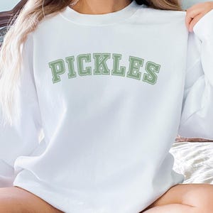 May include: White sweatshirt with the word "PICKLES" printed in green letters with a varsity style font.