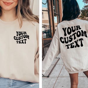 May include: A cream-colored sweatshirt with the text "YOUR CUSTOM TEXT" in black, retro-style lettering. The sweatshirt is shown on a model wearing it both front and back.
