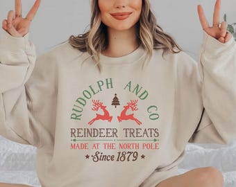 May include: A beige sweatshirt with a festive Christmas design featuring the text "Rudolph and Co Reindeer Treats Made at the North Pole Since 1879". The design includes two red reindeer with green trees and a red star.
