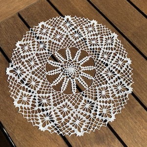 May include: A white, circular, crocheted doily with a floral pattern. The doily is made of delicate, interwoven threads, creating an openwork design. The doily is placed on a wooden surface.