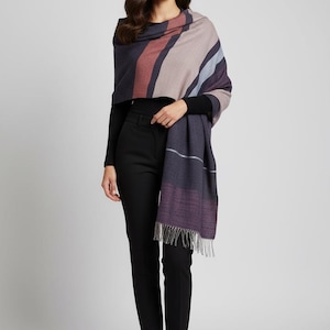 May include: A multi-colored shawl with a striped pattern in shades of purple, red, and gray. The shawl is draped over the shoulders and has a fringed edge. The person is wearing a black long-sleeve top and black pants.