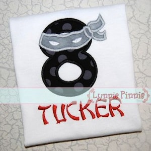 May include: A white t-shirt with a black and white number eight with a gray eye mask and a red headband. The name "Tucker" is embroidered in red below the number.