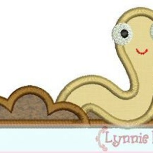 May include: A smiling, light brown worm with large white eyes is embroidered on a brown patch of dirt. The worm is curled into a 'C' shape.