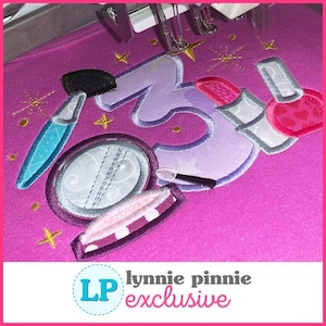 May include: A pink fabric with embroidered makeup-themed designs. The design includes a makeup brush, compact, lipstick, and nail polish, along with the number 3 and starburst accents. The text "lynnie pinnie exclusive" is at the bottom.