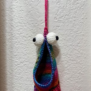 May include: A hanging crochet pouch featuring a vibrant rainbow-coloured yarn body with a wide, open mouth. Two large, round white crocheted eyes with black pupils are attached above the opening. A pink yarn loop extends from the top for hanging.