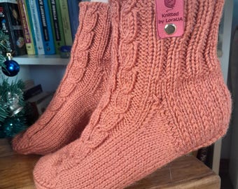 May include: A pair of coral-colored knitted socks with a cable knit design. The socks have a cuff and a tag that reads "Knitted by LoraUA". The socks are displayed on a wooden surface.