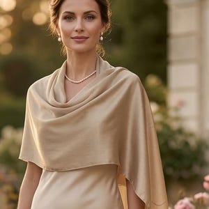 May include: A floor-length champagne-colored satin dress with a matching draped shawl. The dress features a sleeveless bodice with a round neckline and a smooth, flowing skirt that reaches the ground. A simple pearl necklace is worn around the neck.