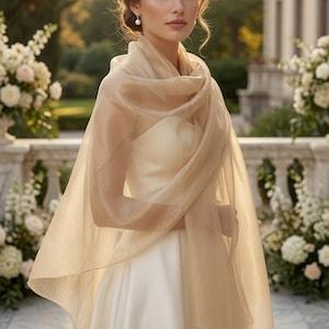 May include: A sheer, champagne-colored shawl draped over the shoulders of a white gown. The lightweight, translucent fabric features a subtle shimmer and soft, flowing edges that extend down the arms, creating a delicate and elegant layered look.