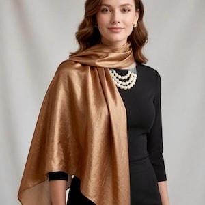 May include: A woman wearing a gold-colored, silky scarf draped over a black dress. The scarf has a subtle sheen and is wrapped around the shoulders. A multi-strand pearl necklace and earrings complete the look. The outfit is suitable for formal occasions.