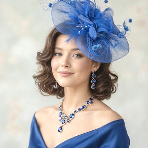 May include: A woman wearing a royal blue hat, earrings, necklace, and a matching off-the-shoulder dress. The hat features a large flower, feathers, and netting. The jewelry has large, blue gemstones. The dress is made of a shiny, satin-like material.