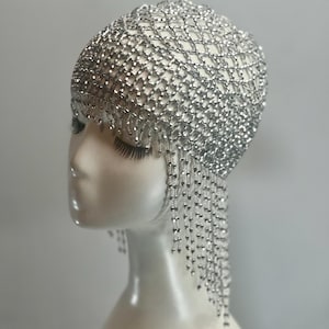 May include: A silver beaded headpiece with a fringe of beads hanging down the back. The headpiece is made of a mesh of small silver beads.