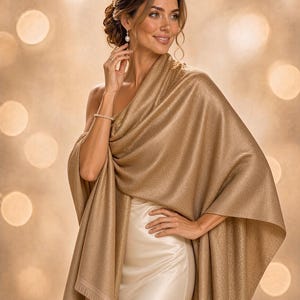 Puede incluir: Un chal dorado brillante drapeado sobre los hombros, con una textura metálica fina y un delicado borde de flecos. La tela tiene una calidad reflectante sutil que capta la luz, creando una apariencia dorada suave y uniforme en toda la prenda.