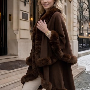May include: A dark brown cape featuring a thick faux fur trim along the collar, front opening, and hemline. The garment is worn over a light beige long-sleeved top and light-colored trousers, creating a layered look with a soft, textured finish.