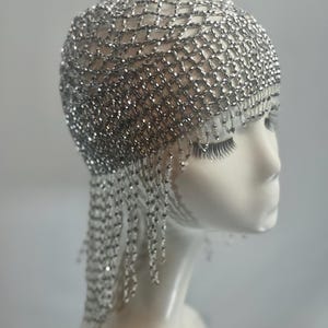 May include: A silver beaded flapper headpiece with a net design and fringe. The headpiece is worn on a white mannequin head.