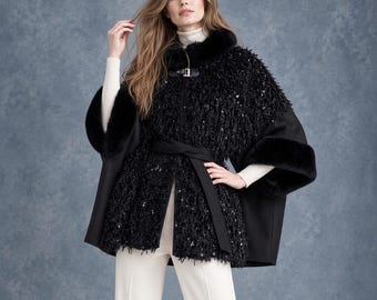 May include: A black cape featuring a textured, sequined front panel, a faux fur collar, and faux fur cuffs. The garment includes a black waist tie and a single buckle closure at the neckline, worn over a cream-colored turtleneck and cream trousers.