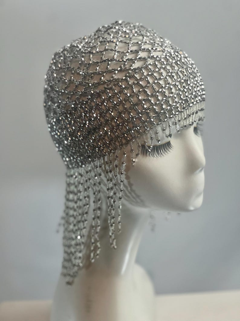 May include: A silver beaded flapper headpiece with a net design and fringe. The headpiece is worn on a white mannequin head.