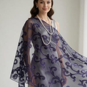 May include: A woman wearing a gray dress and a sheer, dark blue shawl with an ornate, swirling pattern. A long pearl necklace and pearl earrings complete the look. The shawl drapes over the shoulders.