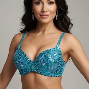 May include: A turquoise bra top featuring dense sequin embellishments and floral-shaped bead accents. The garment includes matching turquoise straps and a structured cup design, fully covered in shimmering blue-green sequins that create a textured surface.