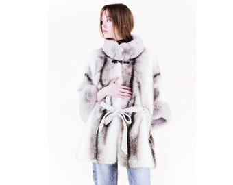 May include: A luxurious, knee-length fur coat in shades of white and gray, featuring a high collar and a matching belt. The coat has a soft, textured appearance and is fastened with a dark clasp. The sleeves have fur cuffs.