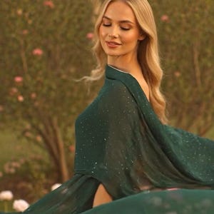 Könnte beinhalten: Ein fließendes smaragdgrünes Kleid mit Umhang, verziert mit dezenten glitzernden Akzenten. Das Kleid hat einen hohen Schlitz und wird im Freien mit einem verschwommenen Hintergrund aus Grün und Blumen getragen.