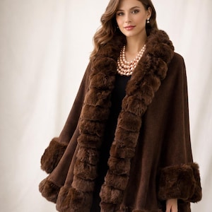 May include: A brown cape with faux fur trim around the collar, front, and cuffs. The cape is a solid color and appears to be made of a soft material. The faux fur is a darker shade of brown, creating a contrast. A multi-strand pearl necklace is also visible.