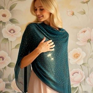 Könnte beinhalten: Ein teal-farbener Schal mit einem zarten Muster aus kleinen, goldfarbenen Punkten. Der Schal ist über den Schultern drapiert. Der Hintergrund zeigt eine florale Tapete mit großen, weichen rosa und weißen Blumen.