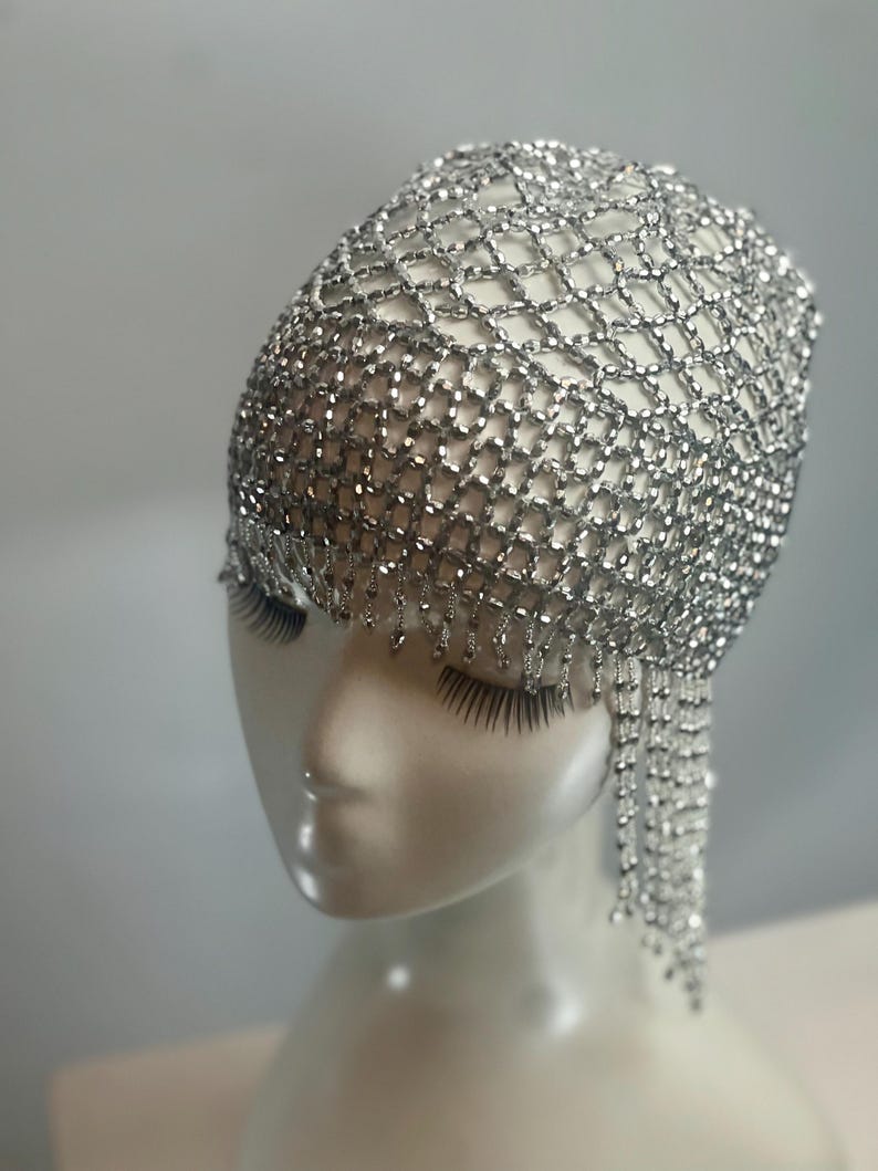 May include: A silver beaded headpiece with a net design and dangling strands. The headpiece is worn on a white mannequin head.