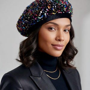 Op de afbeelding: Een zwarte baret bedekt met kleurrijke pailletten. De baret wordt gedragen met een zwarte coltrui, een zwarte blazer en een gouden ketting. De pailletten zijn in verschillende kleuren, waaronder blauw, groen, rood en geel.