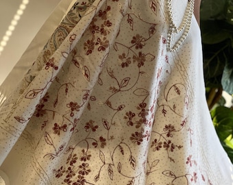 May include: Cream-colored shawl with a floral pattern in shades of brown. The shawl features a delicate design of flowers and leaves, with a dotted border. A pearl necklace is draped over the shawl.