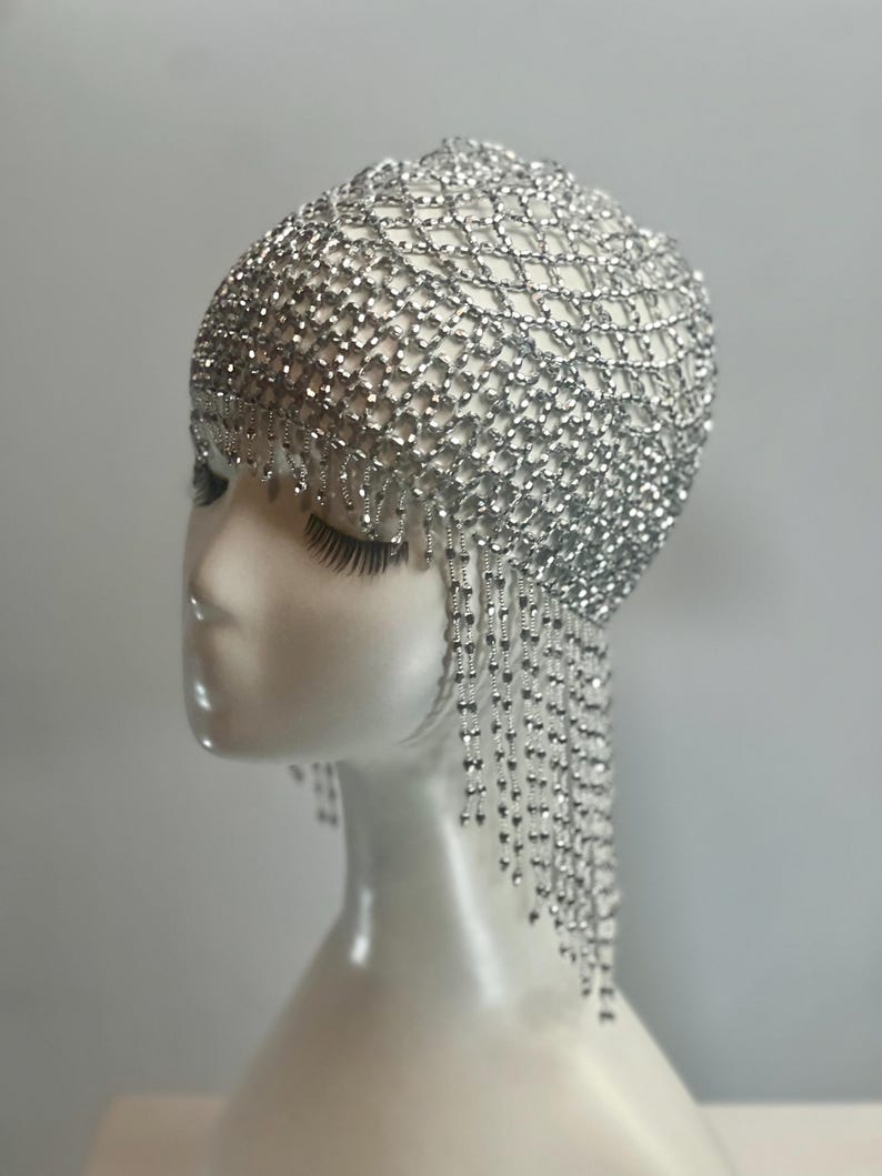 May include: A silver beaded headpiece with a fringe of beads hanging down the back. The headpiece is made of a mesh of small silver beads.