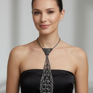 Pode incluir: Um colar de gravata preto com um design decorativo. O colar apresenta uma série de pedras preciosas pretas e uma corrente. O pendente de gravata é longo e tem um padrão intrincado e vazado. A mulher na imagem está usando um top preto sem alças.