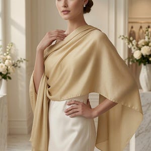 May include: A solid champagne-colored shawl draped over the shoulders, featuring a smooth, lightweight fabric with a subtle sheen. The rectangular wrap is worn over a simple, sleeveless white dress, creating soft folds and elegant drapes.
