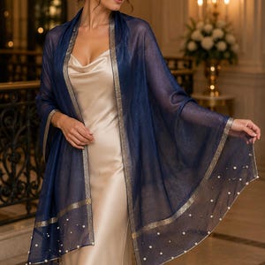 May include: A sheer navy blue shawl featuring a subtle metallic shimmer, gold-toned border trim, and scattered white faux pearl embellishments along the edges. The lightweight wrap is draped over a sleeveless champagne-colored satin slip dress.