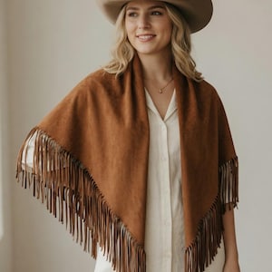 May include: A triangular brown suede shawl featuring long fringe trim along the edges. The shawl is worn over a light cream-coloured button-down shirt. A wide-brimmed tan felt cowboy hat is worn on the head, and a thin gold necklace is visible.