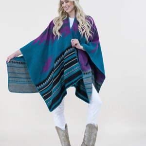 May include: A teal, purple, and black patterned poncho with fringe. The poncho is worn over white jeans and grey cowboy boots.