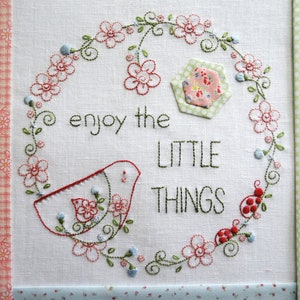 May include: Embroidered white fabric with a green wreath of flowers and leaves, a red bird, and the text "enjoy the little things".