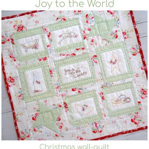 May include: Christmas wall-quilt with embroidered squares featuring holiday motifs. The quilt has a floral pattern with red and pink roses on a white background. The squares are bordered in green and include the text "Joy to the World".