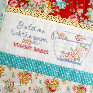 Könnte beinhalten: Ein weißer Stoff-Topflappen mit einer blauen und weißen gestickten Schüssel und dem Text "She let me lick the spoon YUMMY BAKES". Der Topflappen ist aus Patchwork-Stoff mit roten, weißen und blauen Blumenmustern.