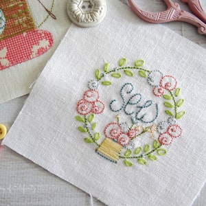 May include: A white fabric square with a hand-stitched embroidery design of a floral wreath with the word "Sew" in the center. The wreath is made of green leaves and pink flowers. A spool of thread is in the center of the wreath.