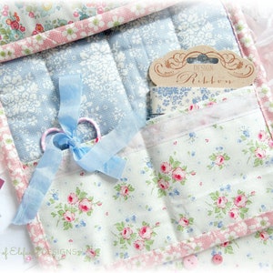 May include: A white fabric storage organizer with multiple pockets, featuring a floral print in shades of pink, blue, and green. The organizer includes a blue ribbon, scissors, and a label that reads "Design Ribbon".