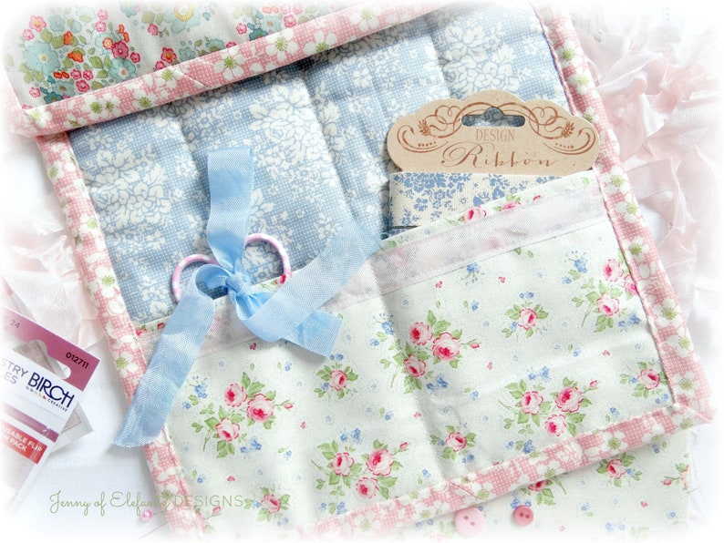 May include: A white fabric storage organizer with multiple pockets, featuring a floral print in shades of pink, blue, and green. The organizer includes a blue ribbon, scissors, and a label that reads "Design Ribbon".