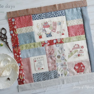 May include: A patchwork quilt block with a simple days theme. The block is made up of various fabrics in shades of red, pink, blue, green, and white. The block features embroidered images of laundry hanging on a line, an iron, and a book with the text "find your joy in the ordinary things".