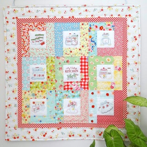 Puede incluir: Una colcha de retazos colorida con un tema de cocina vintage. La colcha presenta varias imágenes bordadas de artículos de cocina, incluyendo frascos, una jarra de galletas, una báscula y un recipiente de harina. La colcha está enmarcada con un borde blanco y tiene un patrón de cuadros rojos y blancos en el centro.
