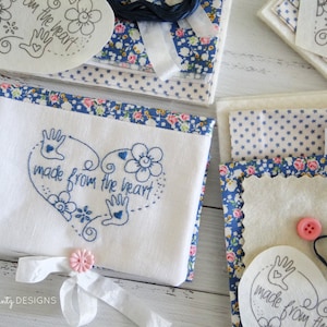 May include: A white embroidery kit with blue floral fabric, white felt, and blue embroidery floss. The embroidery design features a heart with the text "made from the heart" and a handprint. The kit also includes a white ribbon and a pink button.