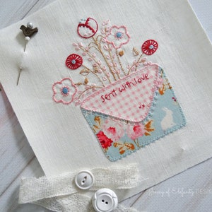 Op de afbeelding: Een wit vierkant van stof met een roze en blauwe envelop met de tekst "Sent with love" erop geborduurd. De envelop is gevuld met roze en rode bloemen en groene stelen. Onderaan de afbeelding zijn twee witte knopen en een stuk wit stof te zien.