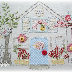 Puede incluir: Bordado que representa una casa con una puerta azul, una ventana con una maceta de flores y un cartel que dice "Bless our home". La casa está rodeada de flores, un árbol con manzanas rojas y una valla de madera.