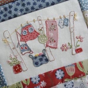 Op de afbeelding: Geborduurd stoffen vierkant met een waslijn met miniatuurkleding, waaronder een rok, handdoeken en een mand. Het ontwerp bevat bloemen- en stippenpatronen in rood, roze en blauw, met geborduurde details en een patchworkrand.