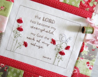 May include: Embroidered white fabric with a quote from Psalm 94:22, "The Lord has become my stronghold, my God the rock of my refuge." The quote is surrounded by a brown frame and a small embroidered bird and flowers.