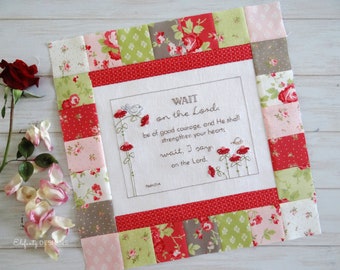 May include: A square, quilted wall hanging with a floral design and the text "WAIT on the Lord." The border is made of red, pink, and green floral fabric squares. Embroidered flowers and a bird accent the text.
