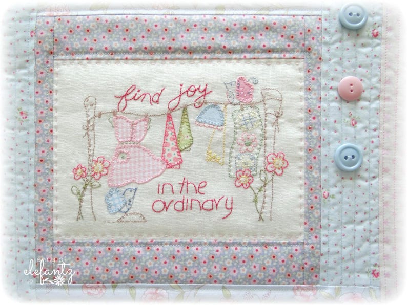 May include: A white fabric wall hanging with pink and blue embroidery. The embroidery depicts a clothesline with clothes hanging on it and the text "find joy in the ordinary".